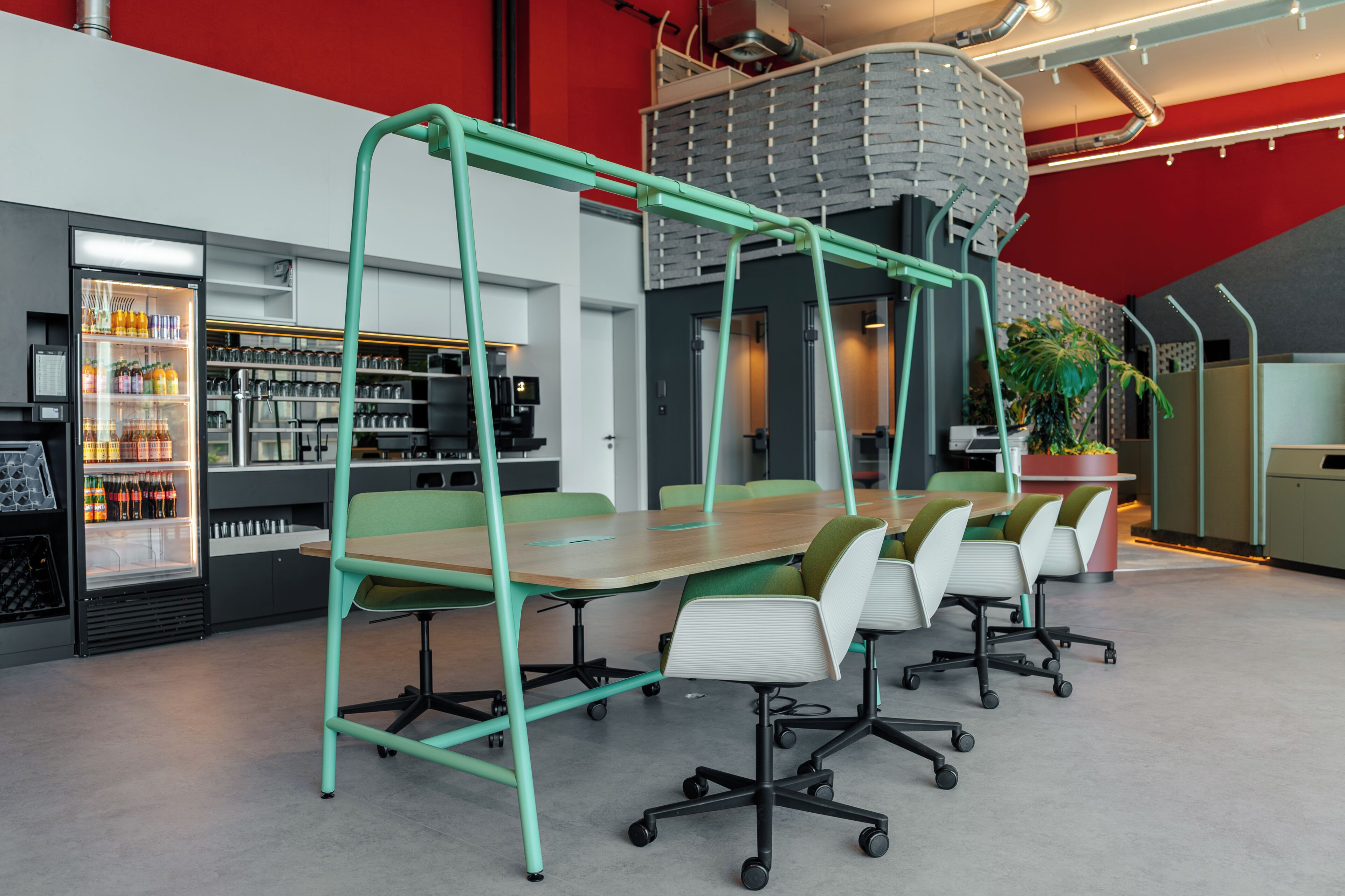 A modern meeting room with a long table, green chairs, and a refrigerator stocked with beverages.