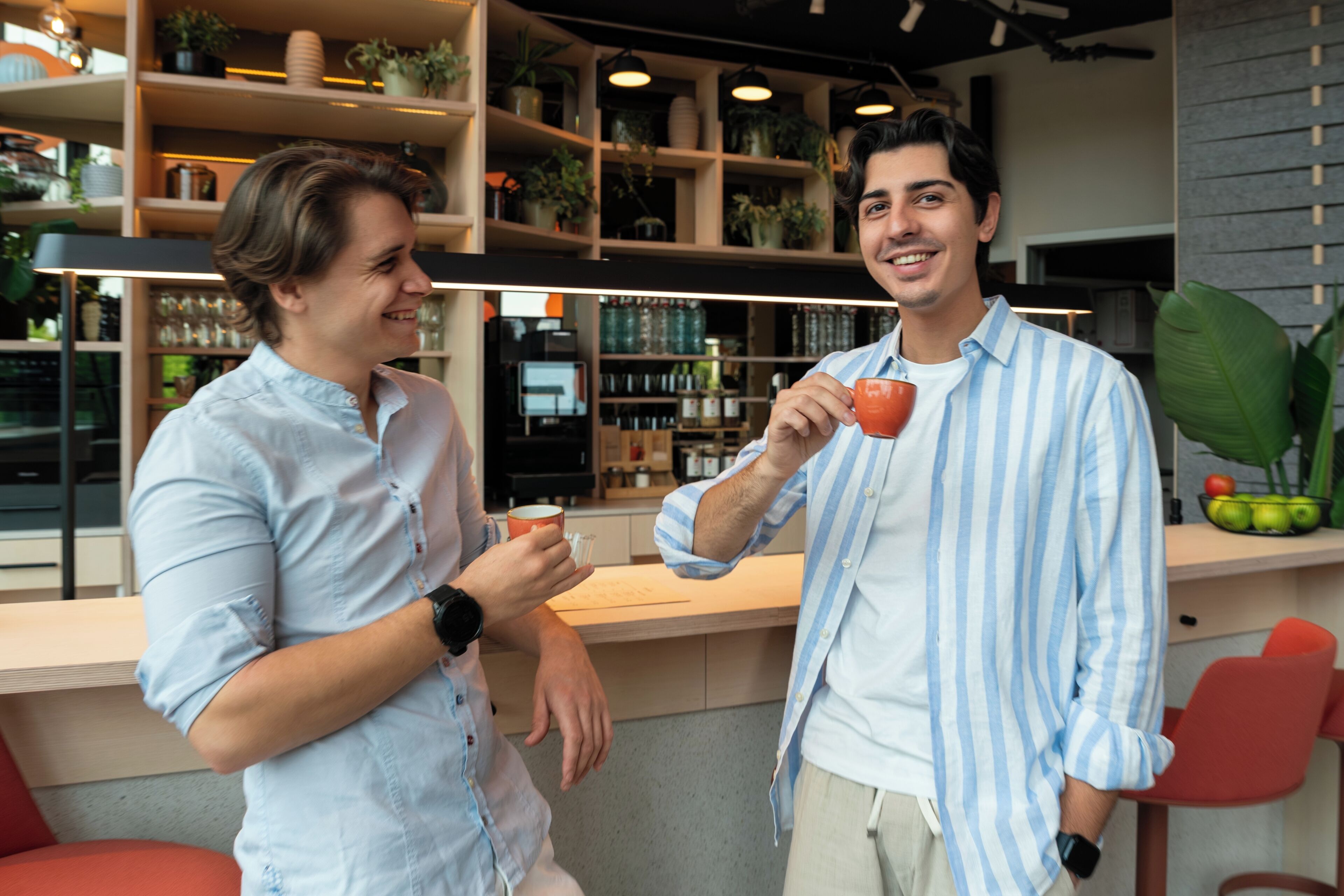 Due uomini in un caffè moderno con scaffali di piante ed elettrodomestici sullo sfondo.