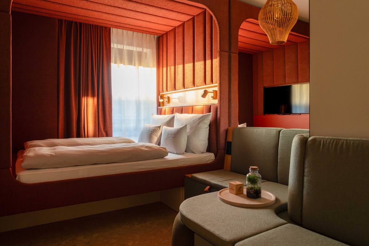 A hotel room with a bed, a sofa, a television, a window with curtains, and a wall-mounted light fixture.