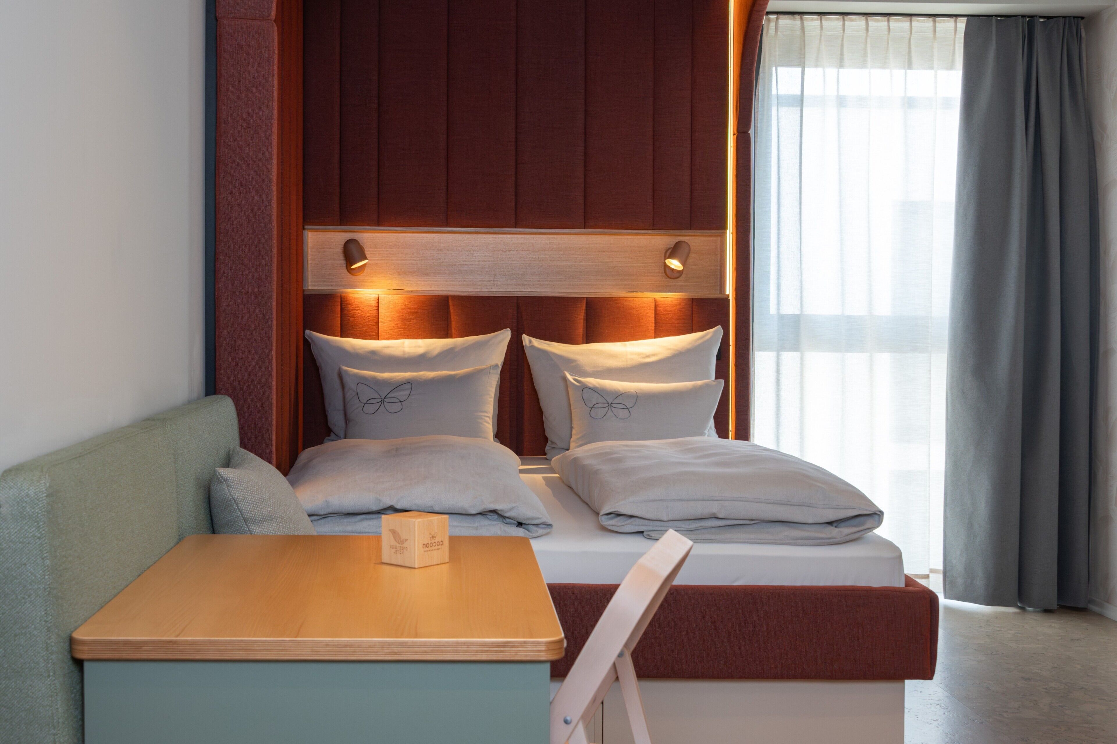 A hotel room with a bed, a wooden nightstand, a seating area, and a window with curtains.