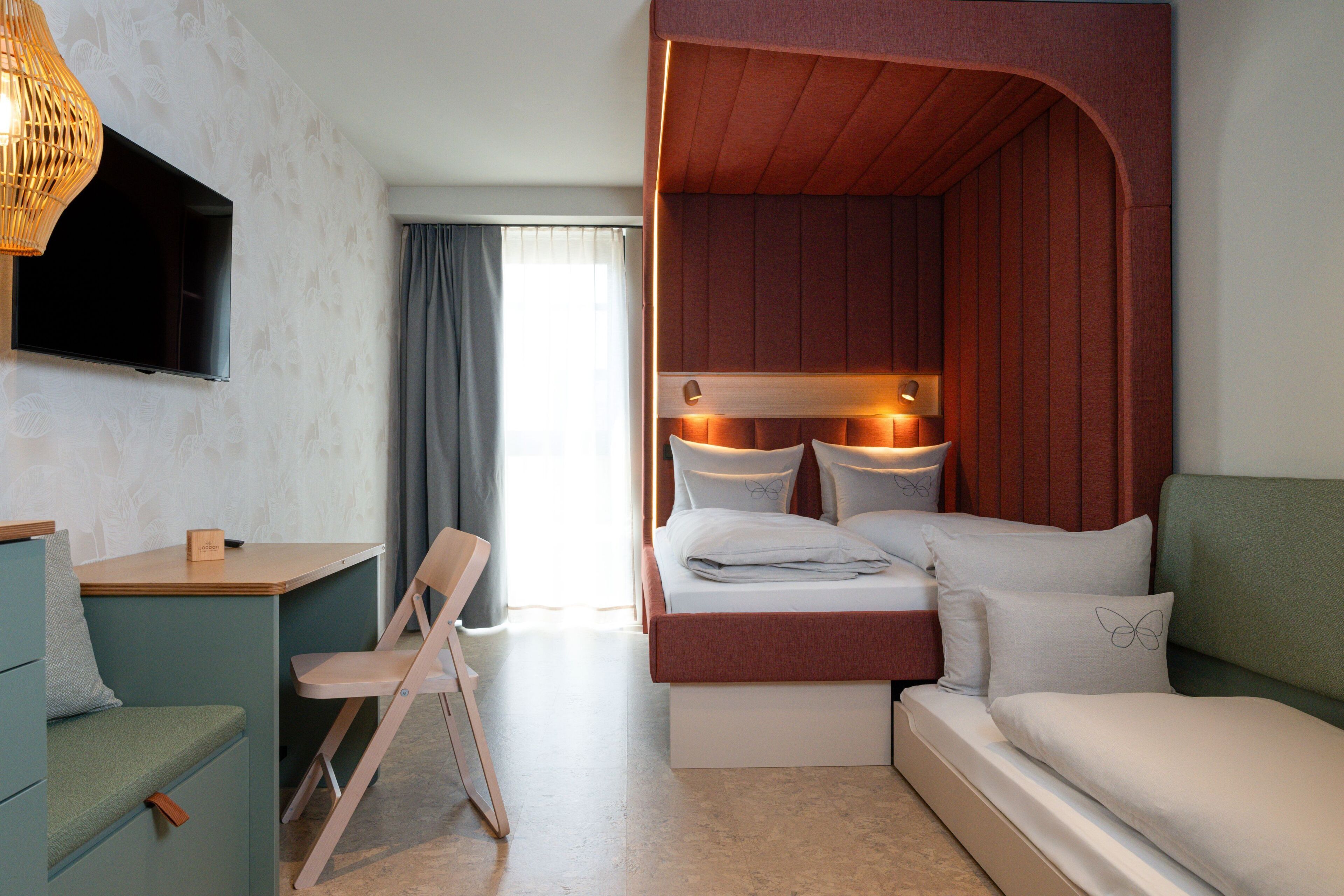 Ein Zimmer mit einem Bett, einem Schreibtisch, einem Sessel, einem Fernseher und einem Fenster mit Vorhängen.