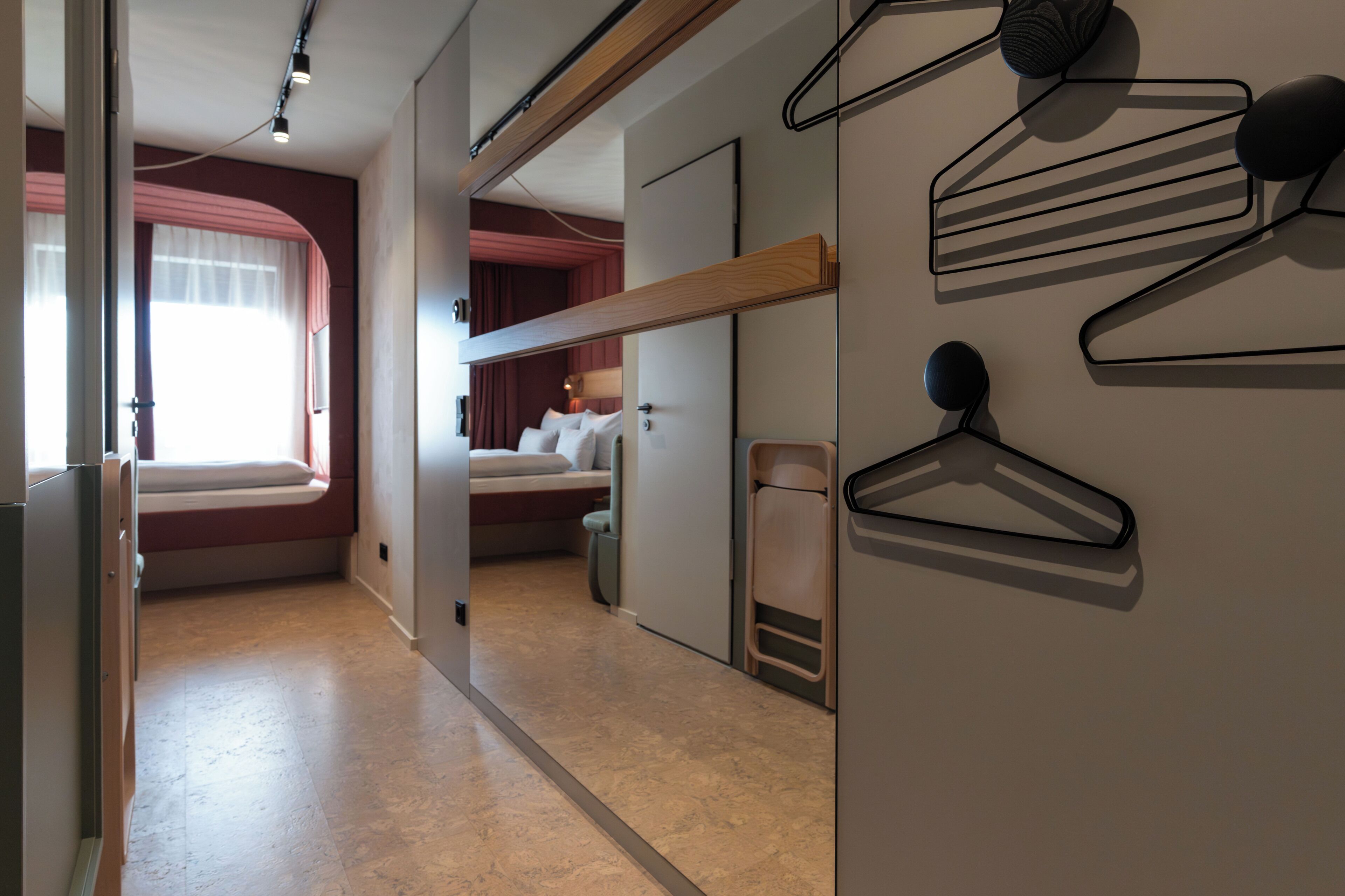 Une chambre d’hôtel avec un lit, une fenêtre avec des rideaux, un porte-manteau fixé au mur et une porte donnant sur une autre pièce.