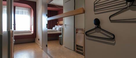 A hotel room with a bed, a window with curtains, a wall-mounted coat rack, and a door leading to another room.