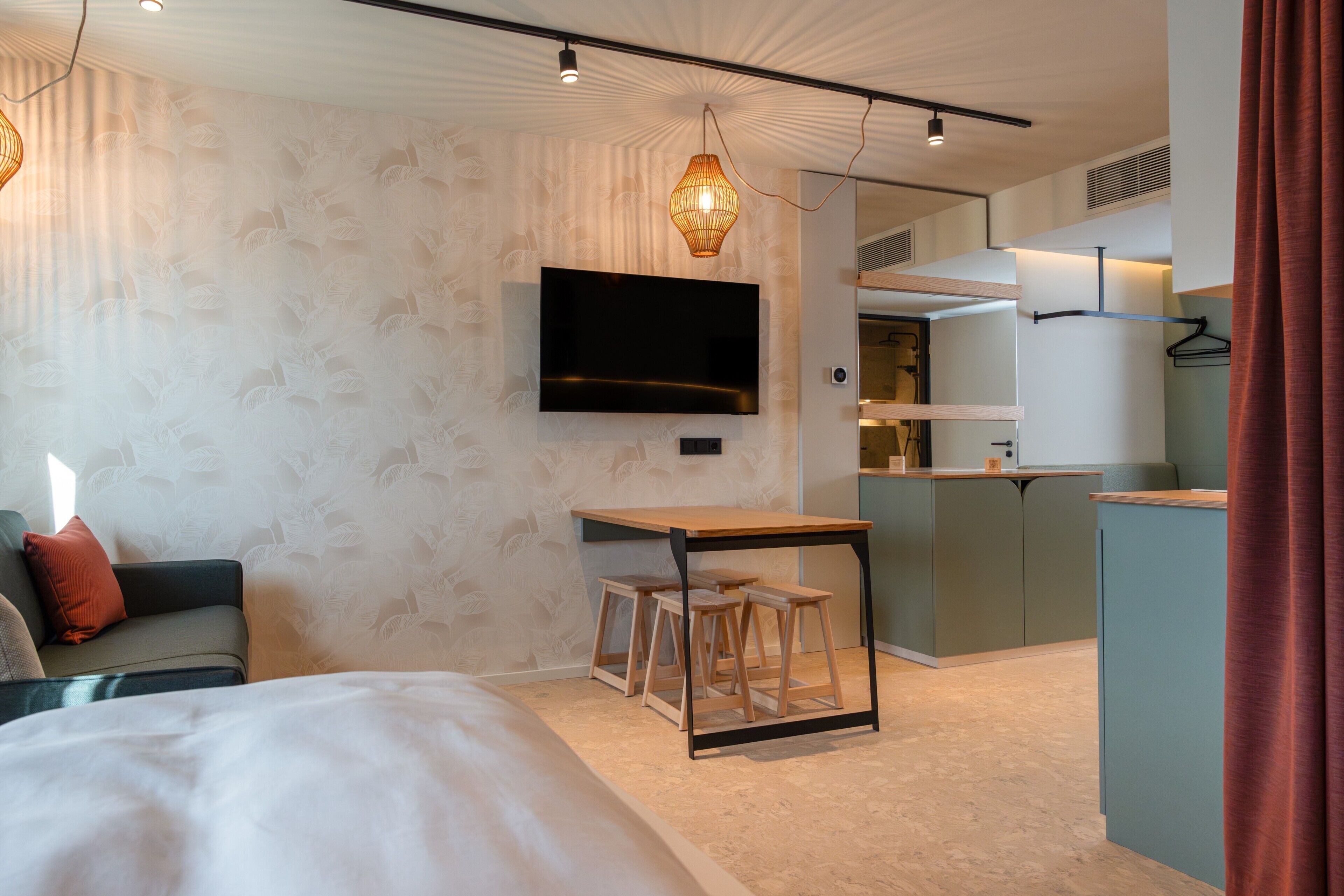 Una habitación de hotel moderna con una cama, un televisor montado en la pared, una pequeña mesa de madera con taburetes y una zona de kitchenette.