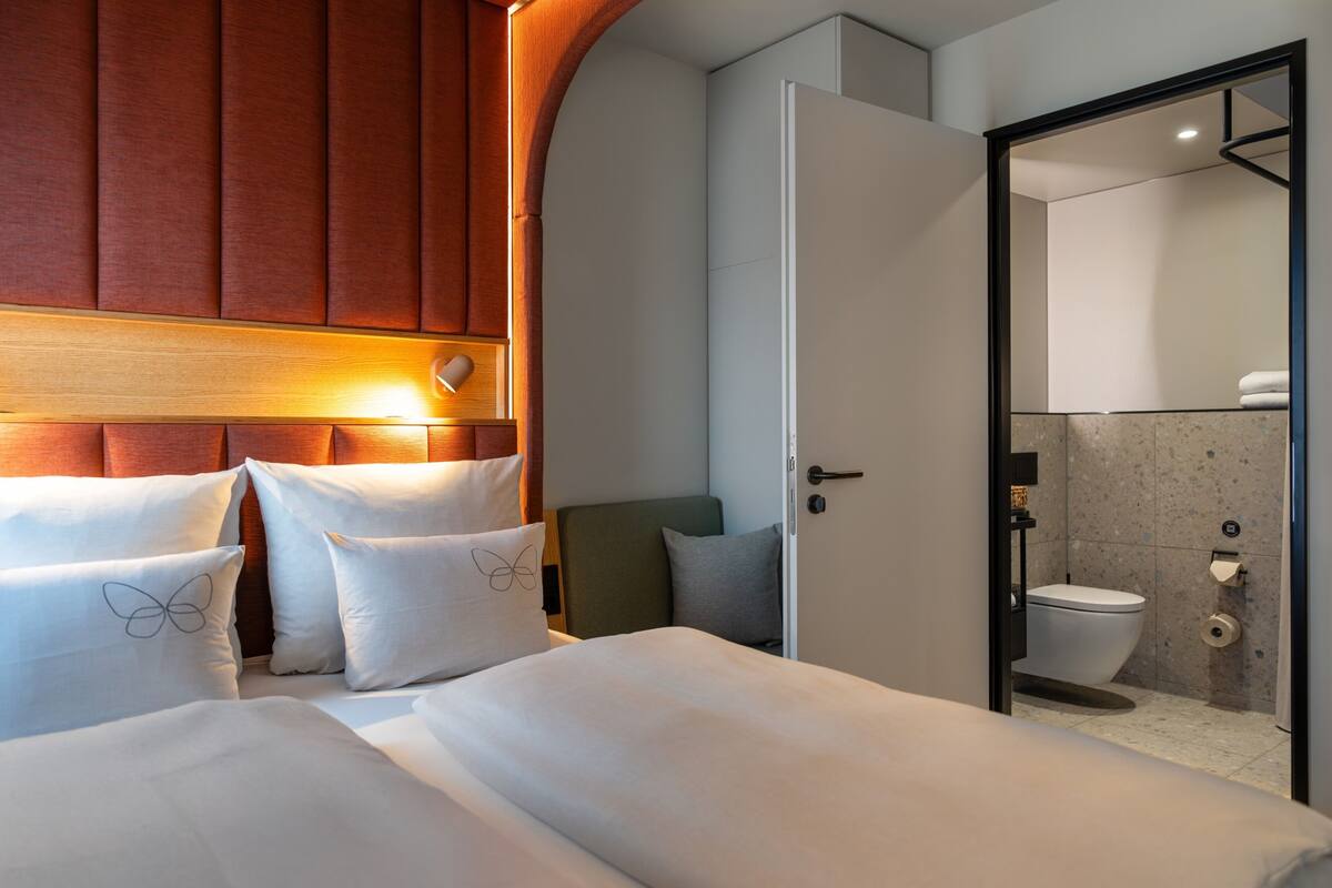 A hotel room with a bed, pillows, a wooden headboard, a door leading to a bathroom with a toilet and shower.