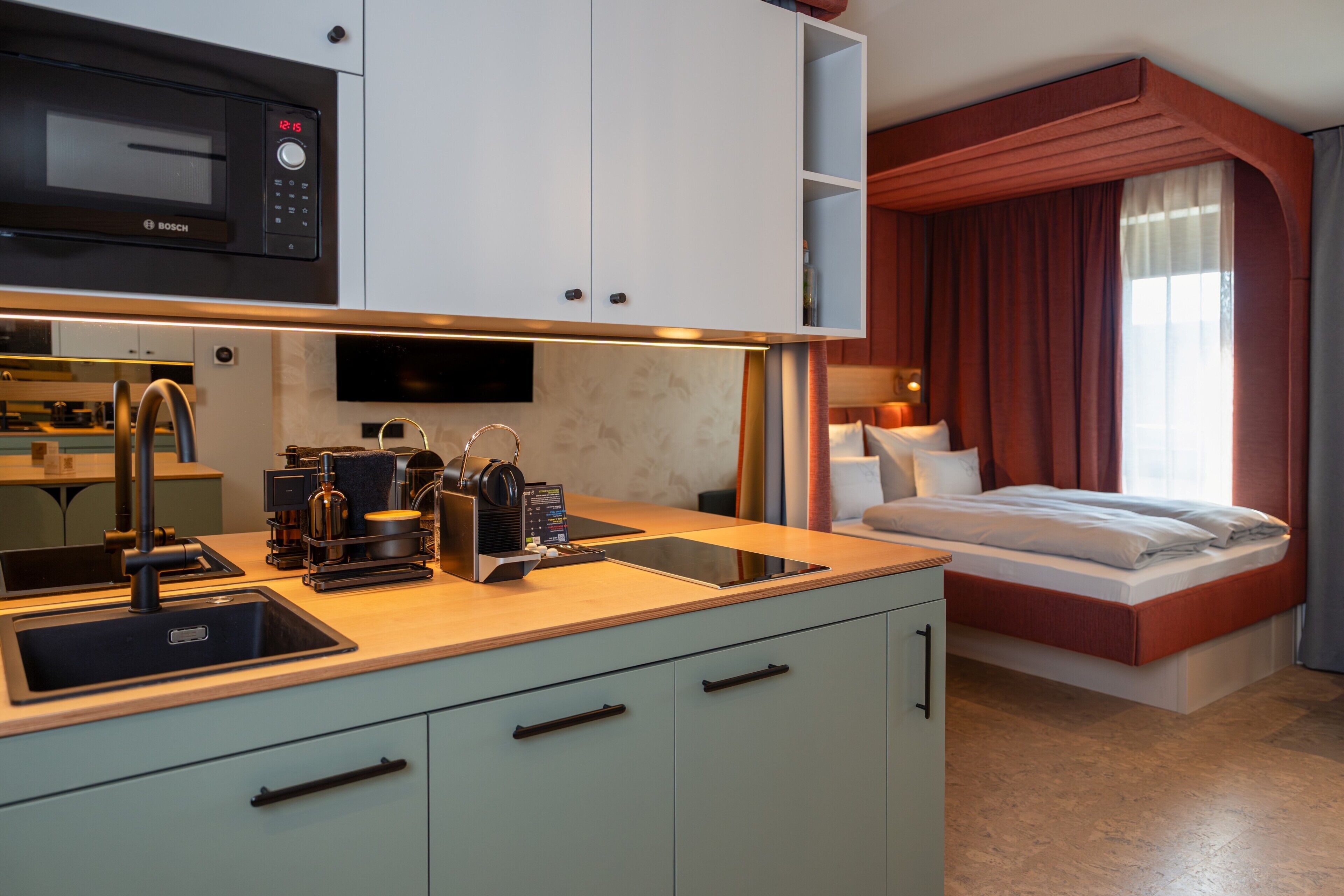 Una habitación de hotel moderna con kitchenette, una cama, un televisor y un ventanal con cortinas.