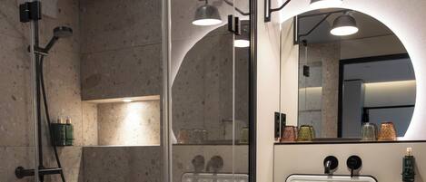 Une salle de bain moderne avec une douche à parois de verre, un lavabo blanc doté d’un robinet noir et un miroir surmonté d’un luminaire circulaire.