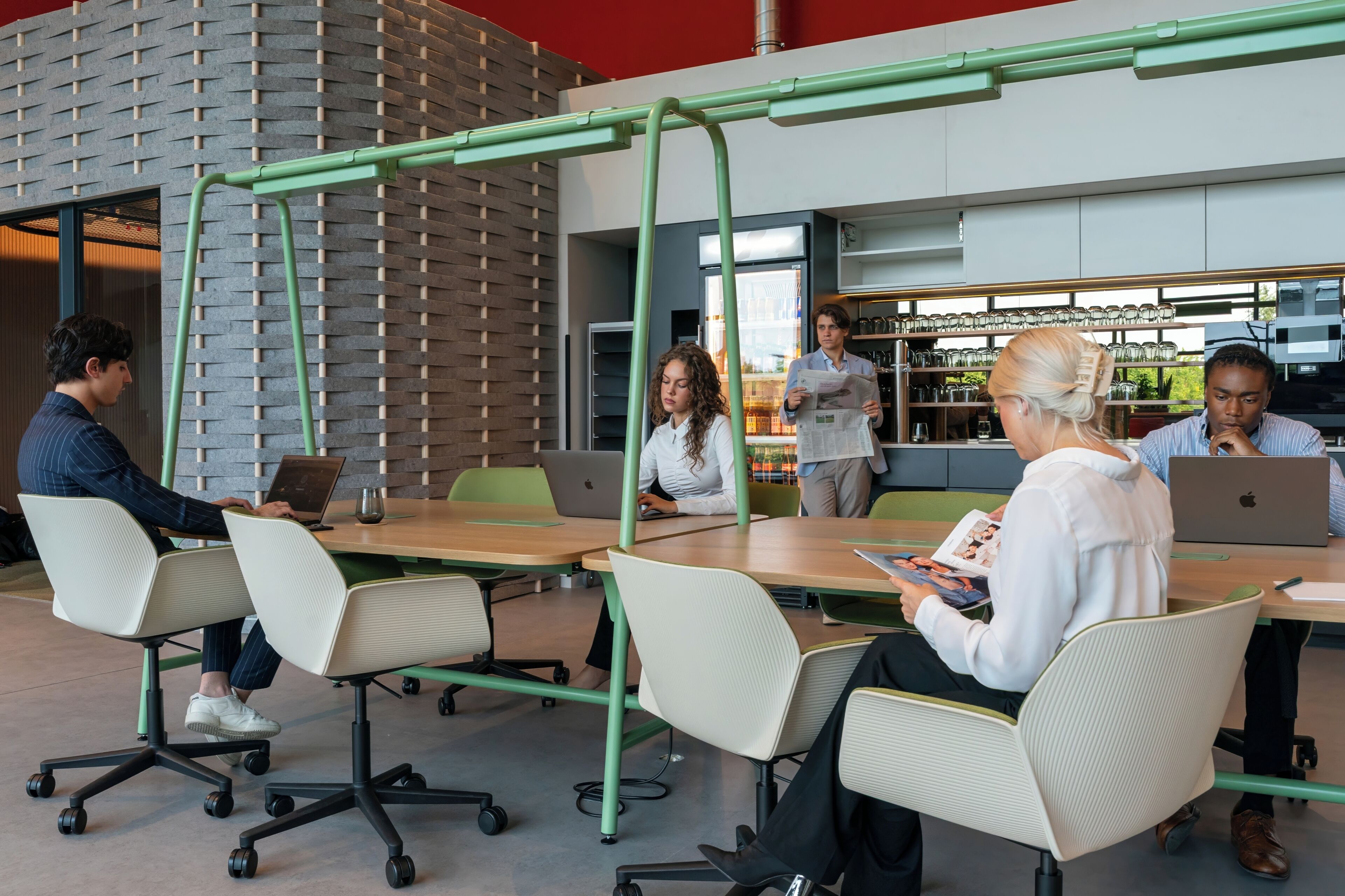 A modern office space with a brick wall, green metal frames, and people working at tables with laptops.