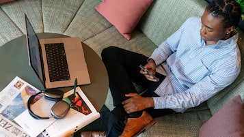 Eine Person sitzt auf einem gemusterten Sofa mit einem Laptop, Kopfhörern und einem Smartphone.