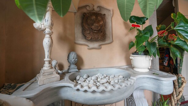 Uma fonte de mármore com cabeça de leão, rodeada por plantas e um vaso branco.