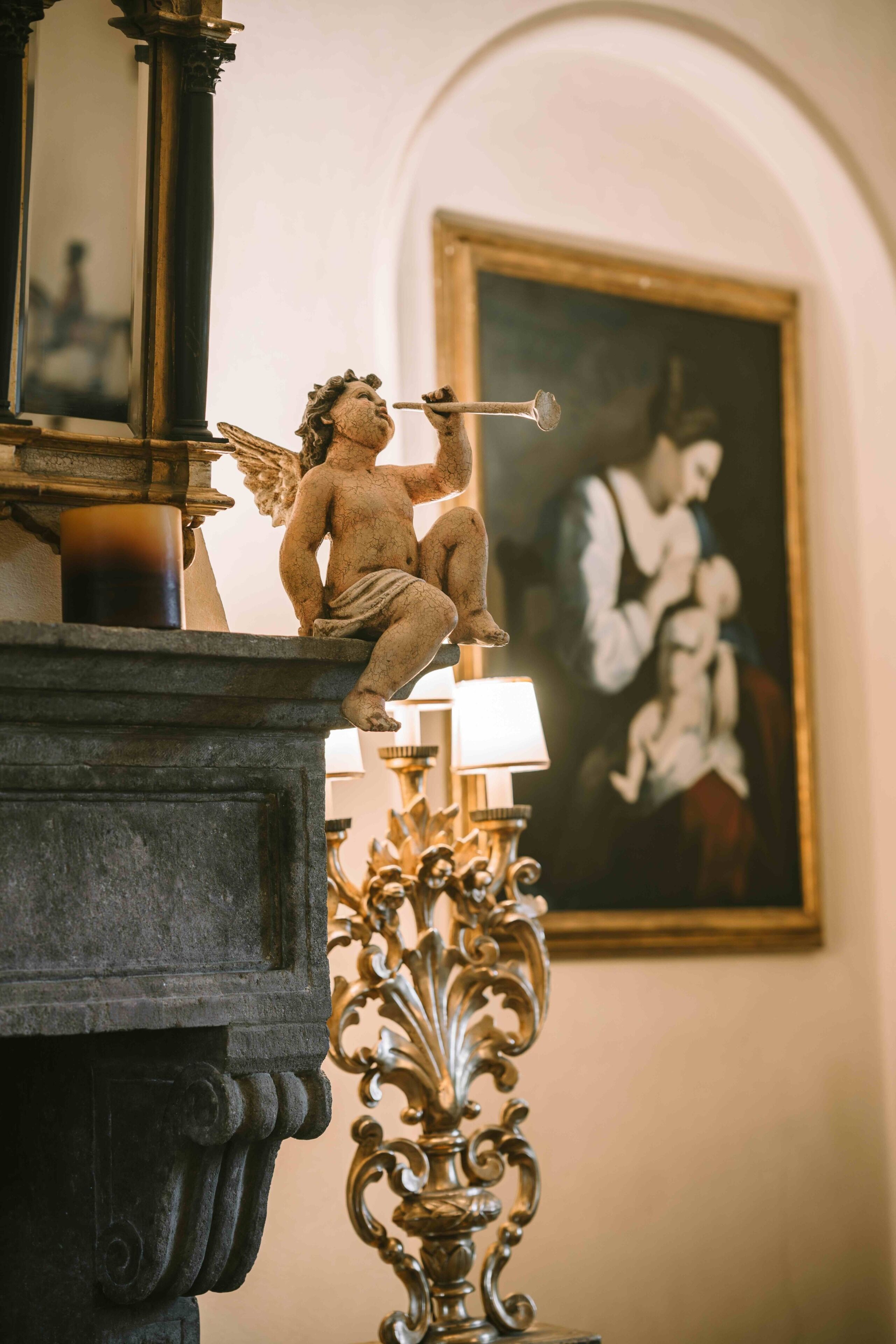 A statue of a cherub on a mantelpiece, a framed painting on the wall, and an ornate lamp.