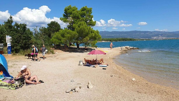 Una playa con gente relajándose, una tienda de campaña y una vista despejada del mar y las montañas al fondo.