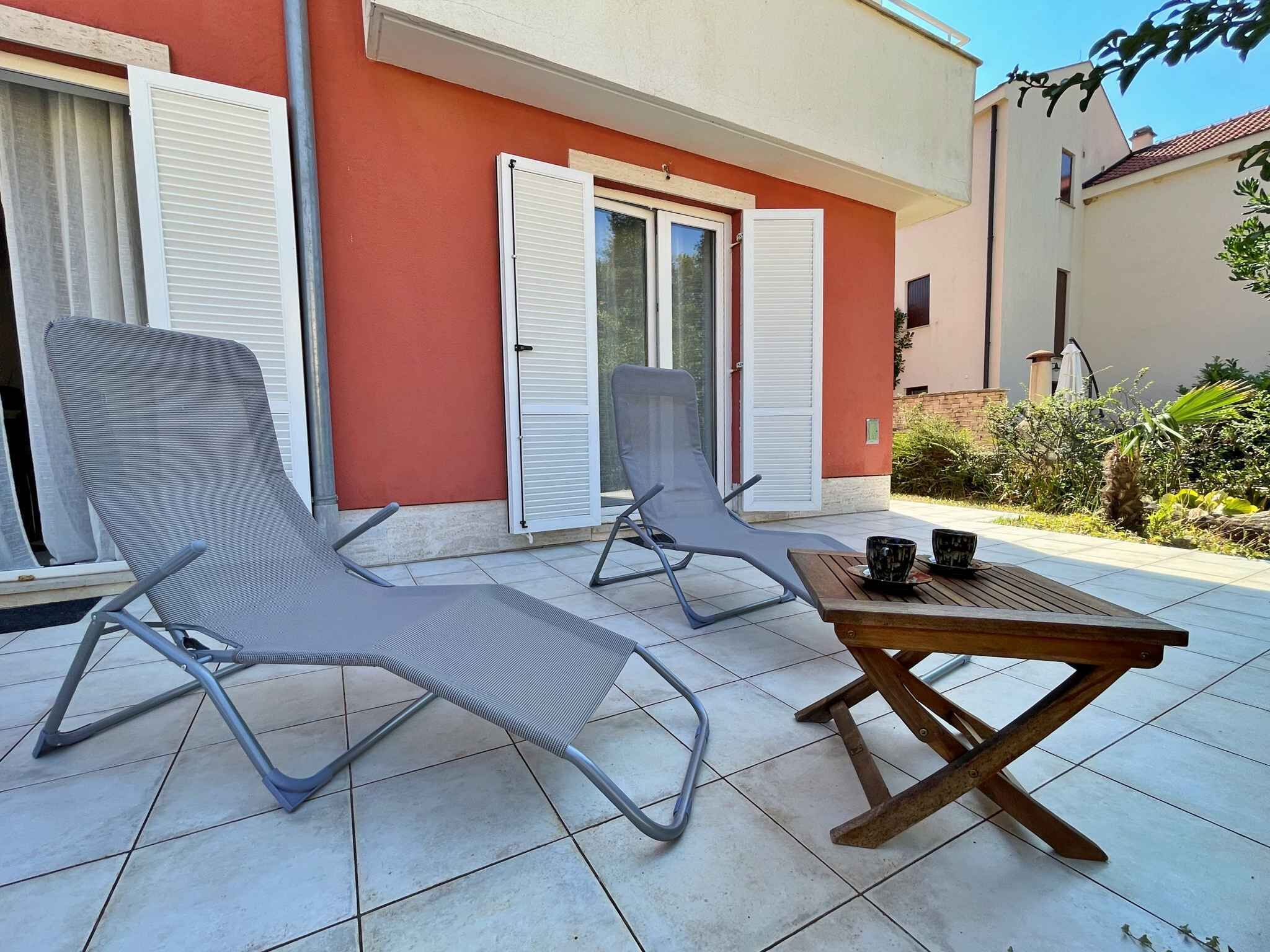 Twee lounge stoelen en een klein houten tafeltje op een betegeld terras, met een rood gebouw op de achtergrond.
