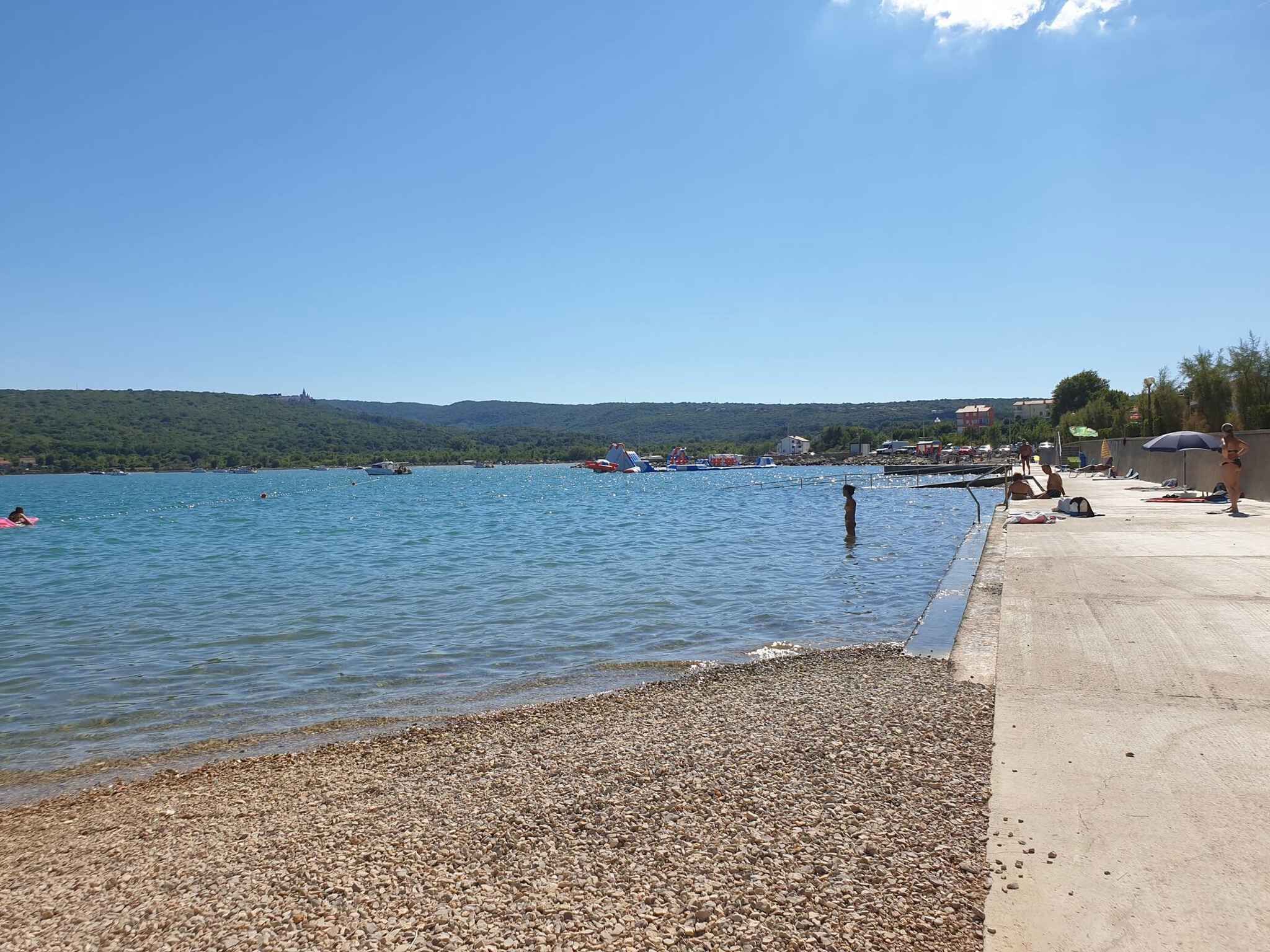 Ein Kiesstrand mit blauem Himmel, ruhigem Wasser und Booten in der Ferne.