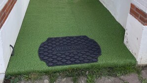 Un espacio exterior cubierto con césped artificial, una alfombra de bienvenida y una puerta de madera.