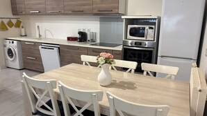 Una cocina moderna con armarios de madera, horno empotrado, microondas y lavavajillas. Hay una mesa de comedor con sillas y un jarrón con flores.