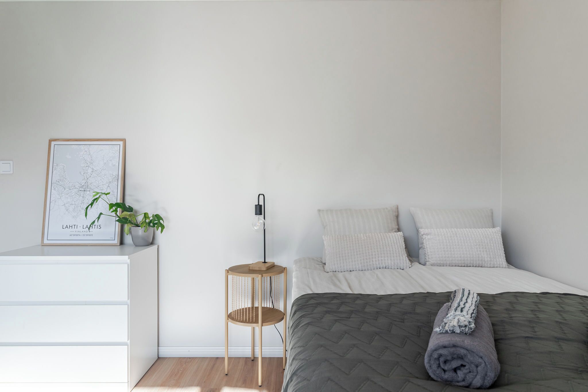 A minimalist bedroom with a bed, a bedside table, a small wooden stool, a framed map, and a potted plant.