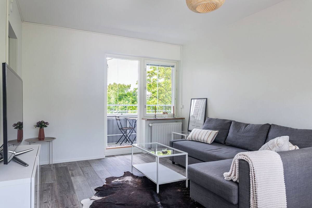 Un salon moderne avec un canapé gris, une table basse en verre et un tapis noir et blanc. On y trouve également un radiateur blanc, une petite table avec un vase et un tableau encadré accroché au mur.
