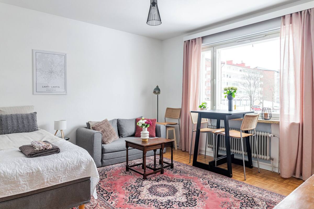Une chambre à coucher moderne avec un lit, un canapé gris, une petite table avec un vase et une fenêtre avec des rideaux.