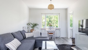 A modern living room with a grey sofa, a glass coffee table, a dining area with a white table and chairs, and a large window offering a view of greenery.