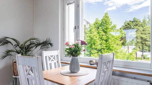 A dining area with a wooden table, white chairs, a vase with flowers, and a view of greenery through a window.
