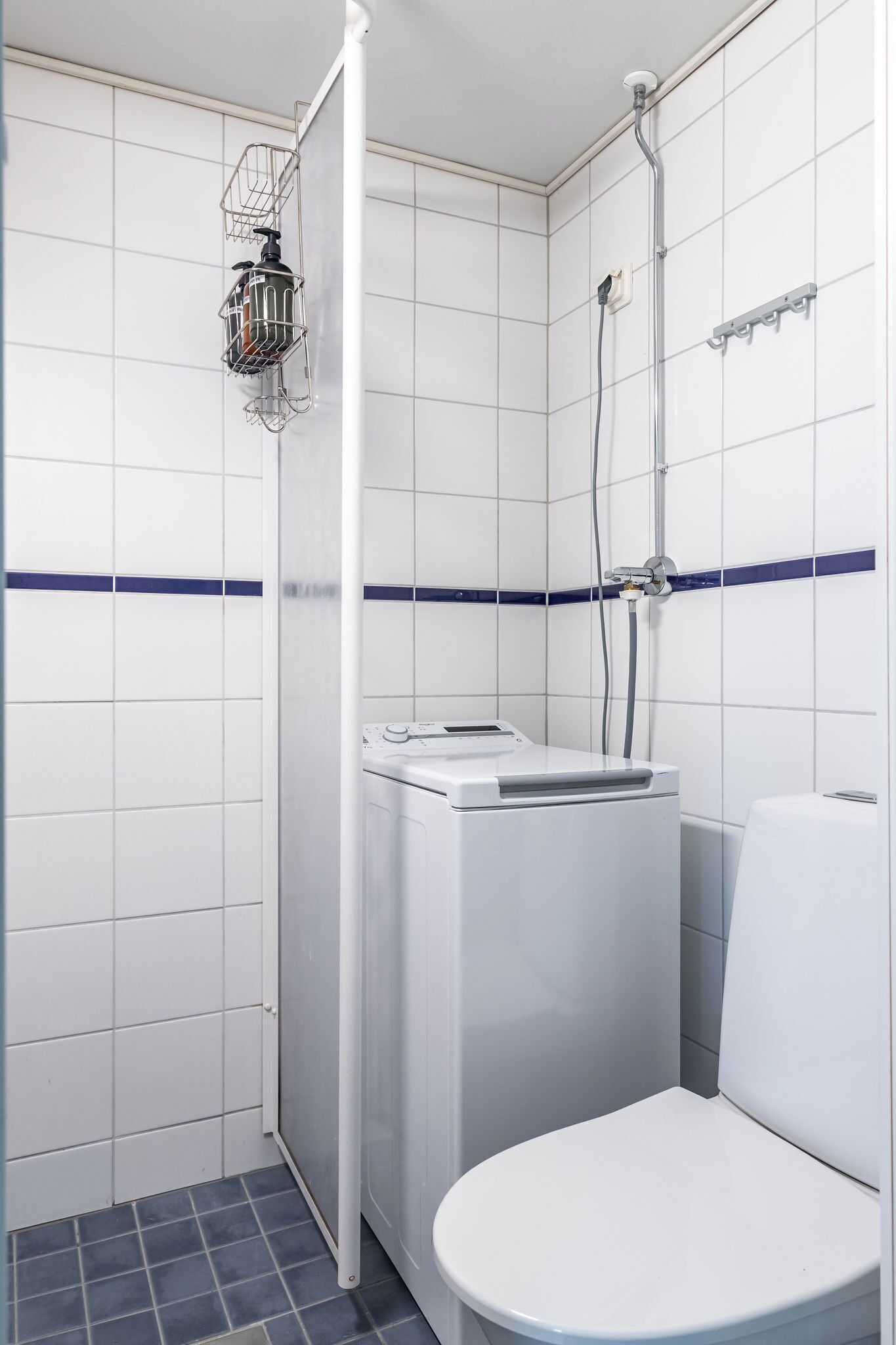 Et lille, rent badeværelse med hvide fliser, en bruser, toilet og vaskemaskine.