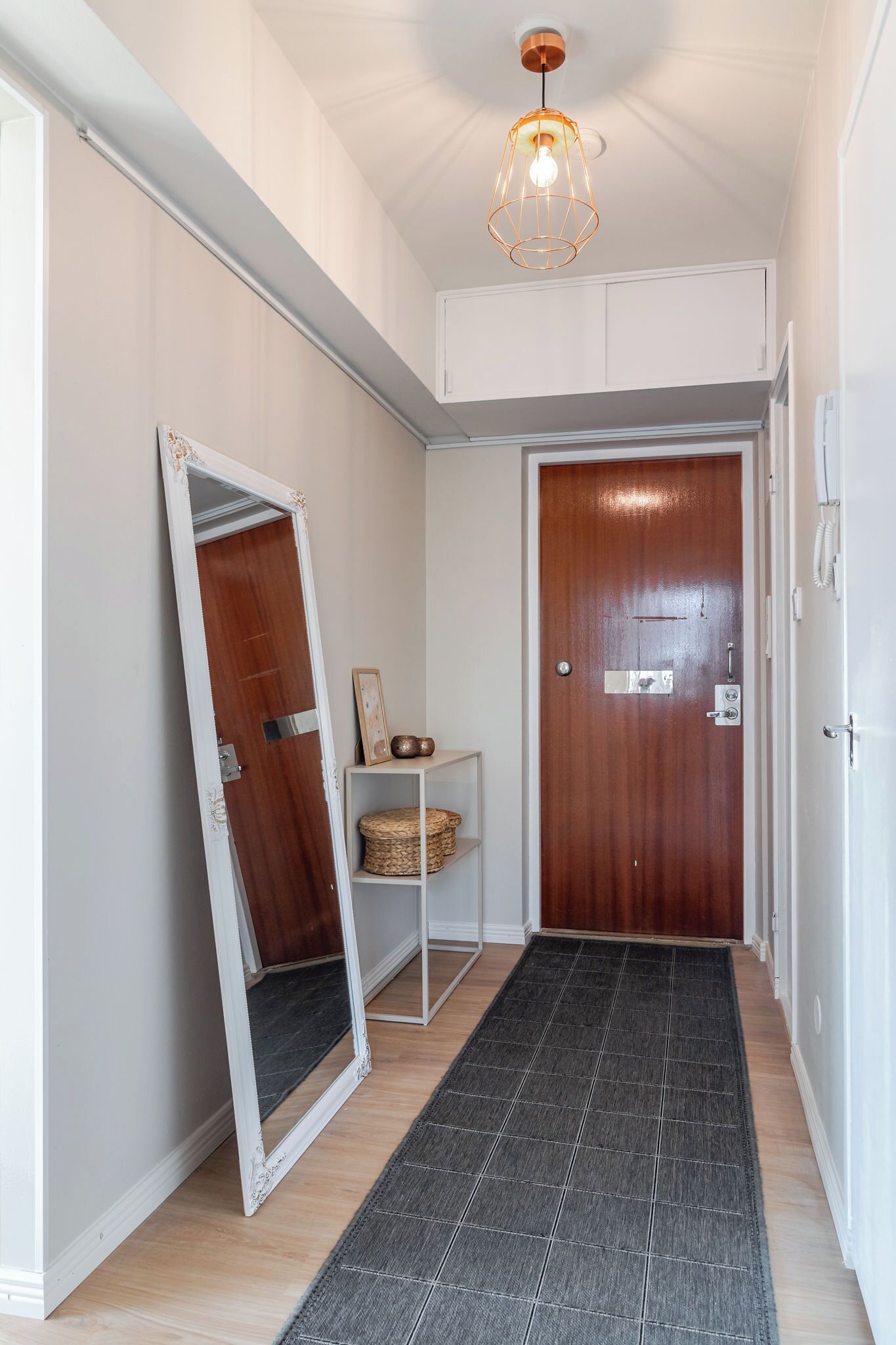 Un pasillo con una puerta de madera, un espejo de marco blanco apoyado en la pared, una pequeña estantería con objetos decorativos y una alfombra gris oscuro en el suelo.