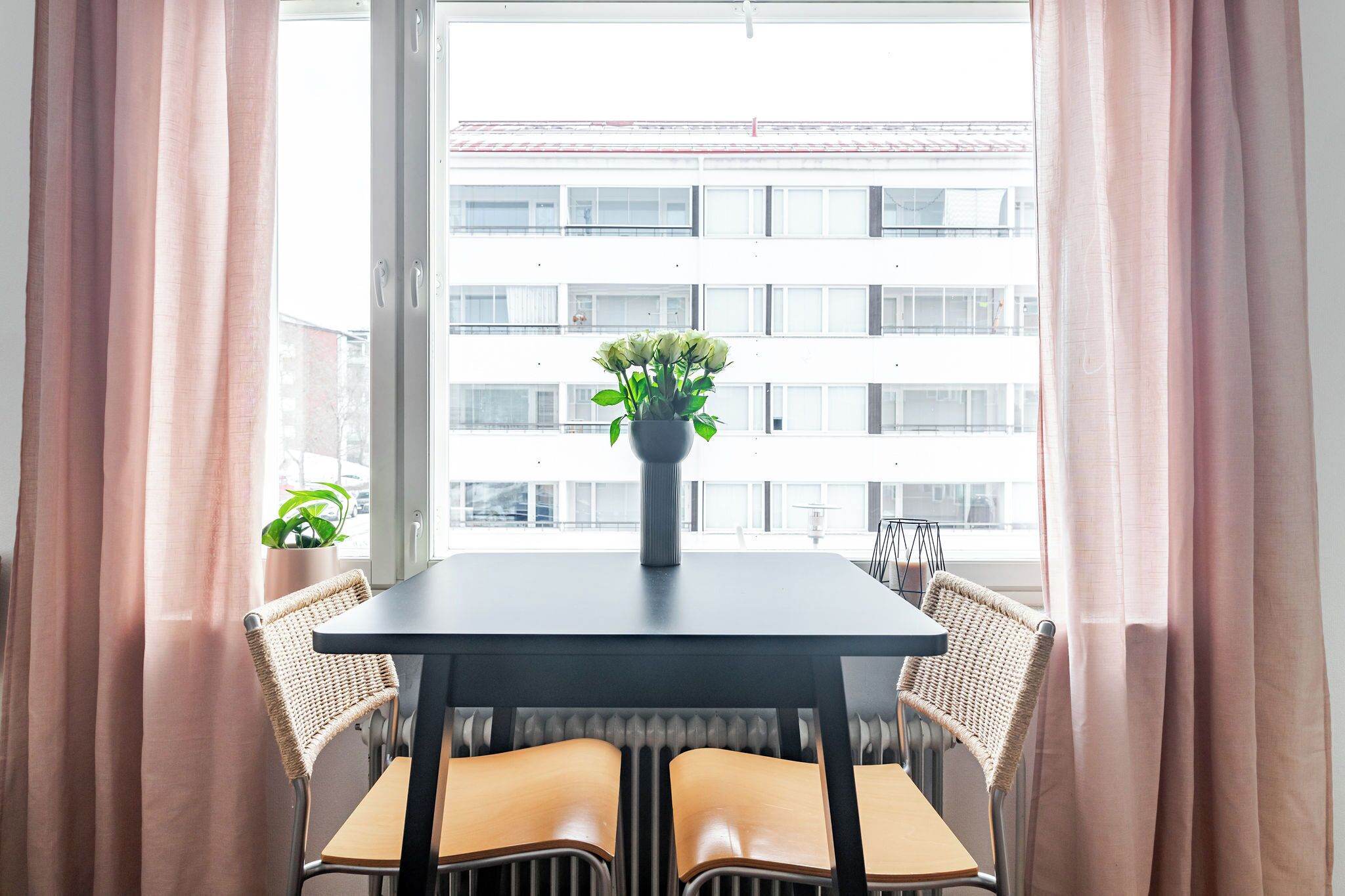 Zona pranzo con tavolo e sedie, una pianta in vaso sul tavolo e vista su un edificio attraverso una finestra con tende.