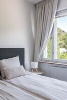 Un dormitorio con cama, mesita de noche, lámpara y ventana con cortinas.