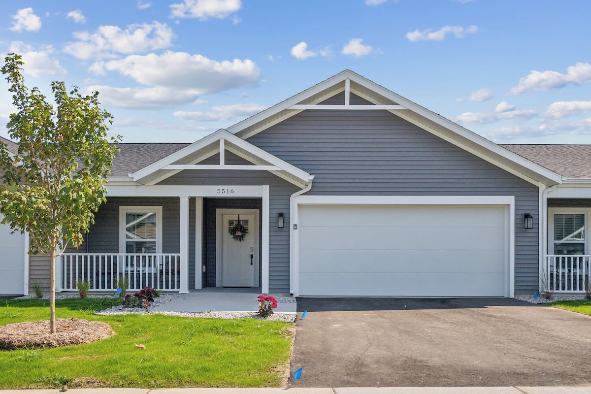 A single-story house with a garage, a front porch, and a small landscaped area with a tree and flowers.