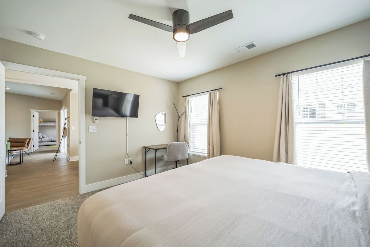A bedroom with a large bed, a ceiling fan, a TV mounted on the wall, and a window with curtains.