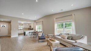 A spacious living area with a sofa, coffee table, and a dining table with chairs. There is a kitchen in the background with cabinets and a window. - Traverse City Flats (Williamsburg)