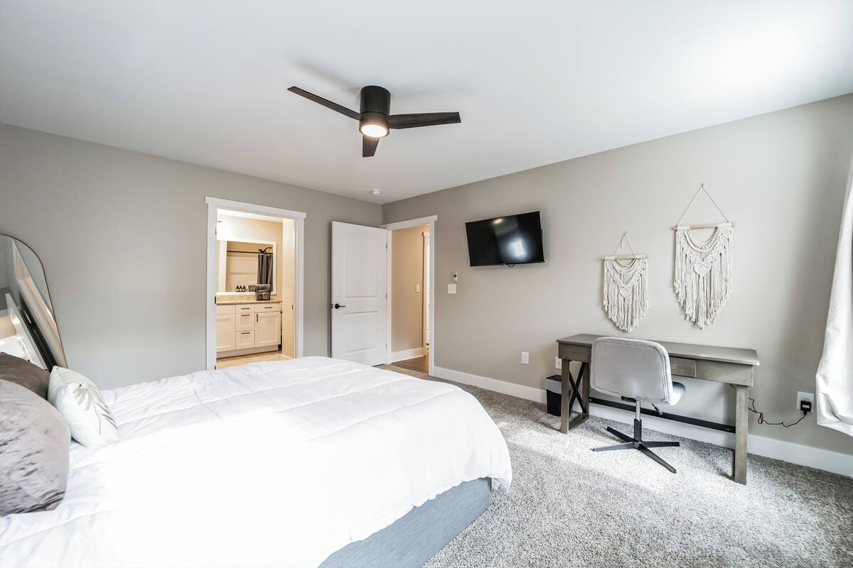 A bedroom with a bed, a desk, a chair, a ceiling fan, a TV, and macrame wall decor.