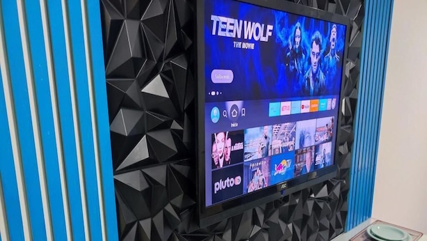 Een moderne kamer met een tv waarop 'TEEN WOLF DE FILM' via Pluto TV wordt vertoond, een lange tafel met borden en bestek, en een geometrisch wandontwerp.