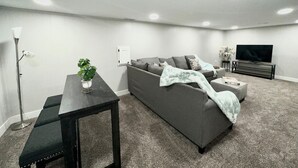 A modern living room with a grey sofa, a small table with a plant, a TV, and a carpeted floor. - Central 5-BR Retreat with Basement Kitchenette & Convenient Freeway Access (Westminster)