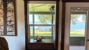 Une pièce comprenant un bureau en bois, une chaise, un canapé en cuir marron et une grande fenêtre donnant sur un lac et sur du mobilier d’extérieur.