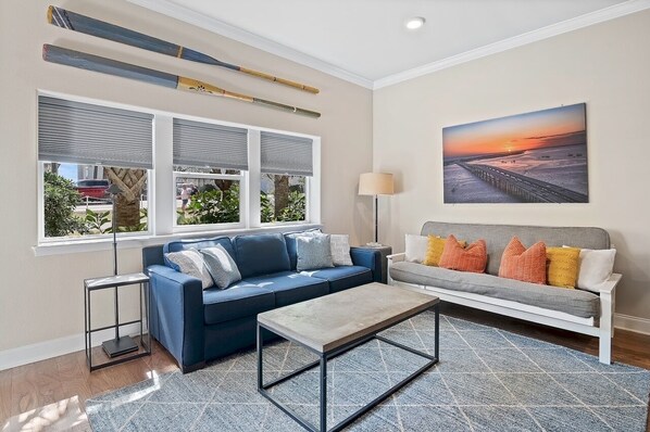 Un salon moderne avec un canapé bleu, un canapé gris agrémenté de coussins orange, une table basse et un tableau représentant un coucher de soleil.