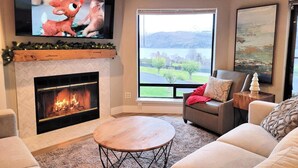 A living room with a fireplace, a television mounted above it, a large window with a view of a lake and hills, and a cozy seating area with a sofa and a coffee table.