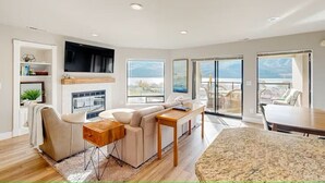 A modern living room with a fireplace, a sofa, a dining table, and large windows offering a view of the outdoors.