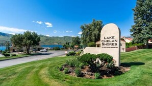 A sign for Lake Chelan Shores with a view of a residential area and mountains in the background.