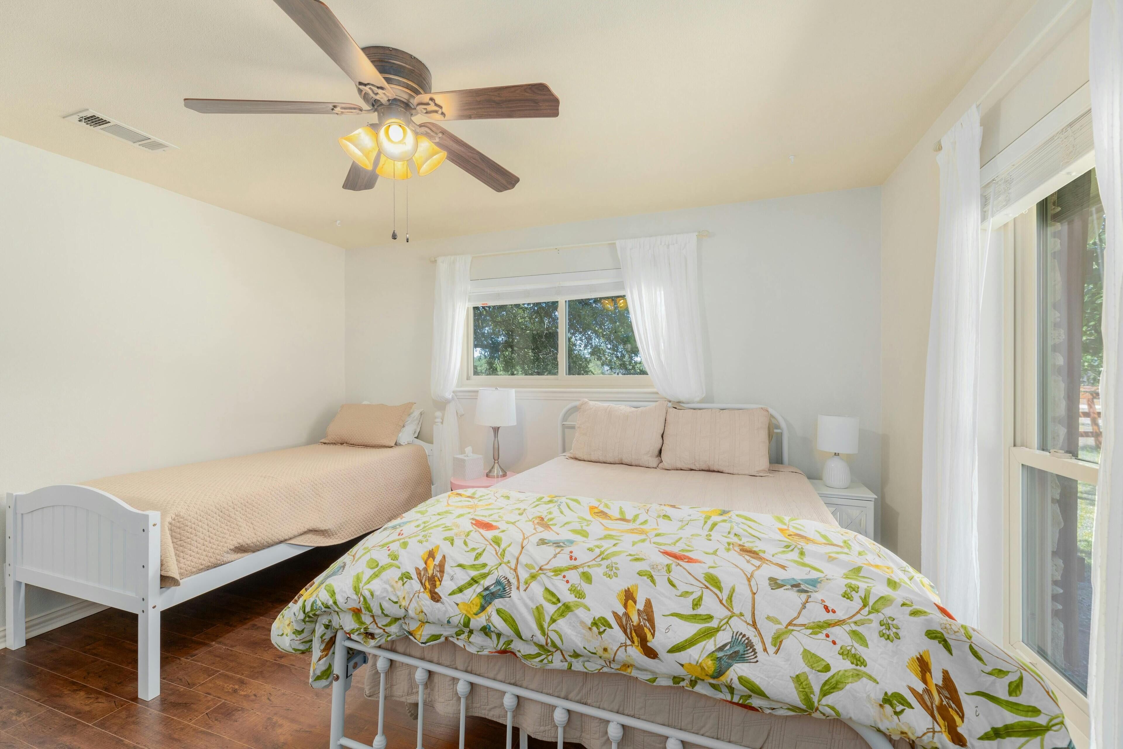 Une chambre avec deux lits simples, un ventilateur de plafond, une fenêtre avec des rideaux et une vue sur la verdure.