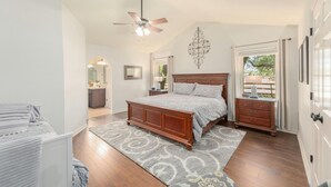 A bedroom with a wooden bed, a ceiling fan, a nightstand, a dresser, and a large window with curtains. - Beautiful Pool home on 2.5 acres with a serene country feel with urban location (Round Rock)