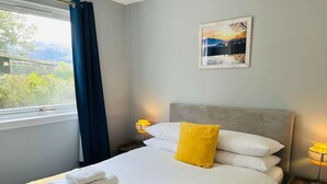 Ein ordentlich bezogenes Bett mit weißen Leinen und einem gelben Kissen, ein Nachttisch mit einer Lampe und ein Fenster mit blauen Vorhängen.