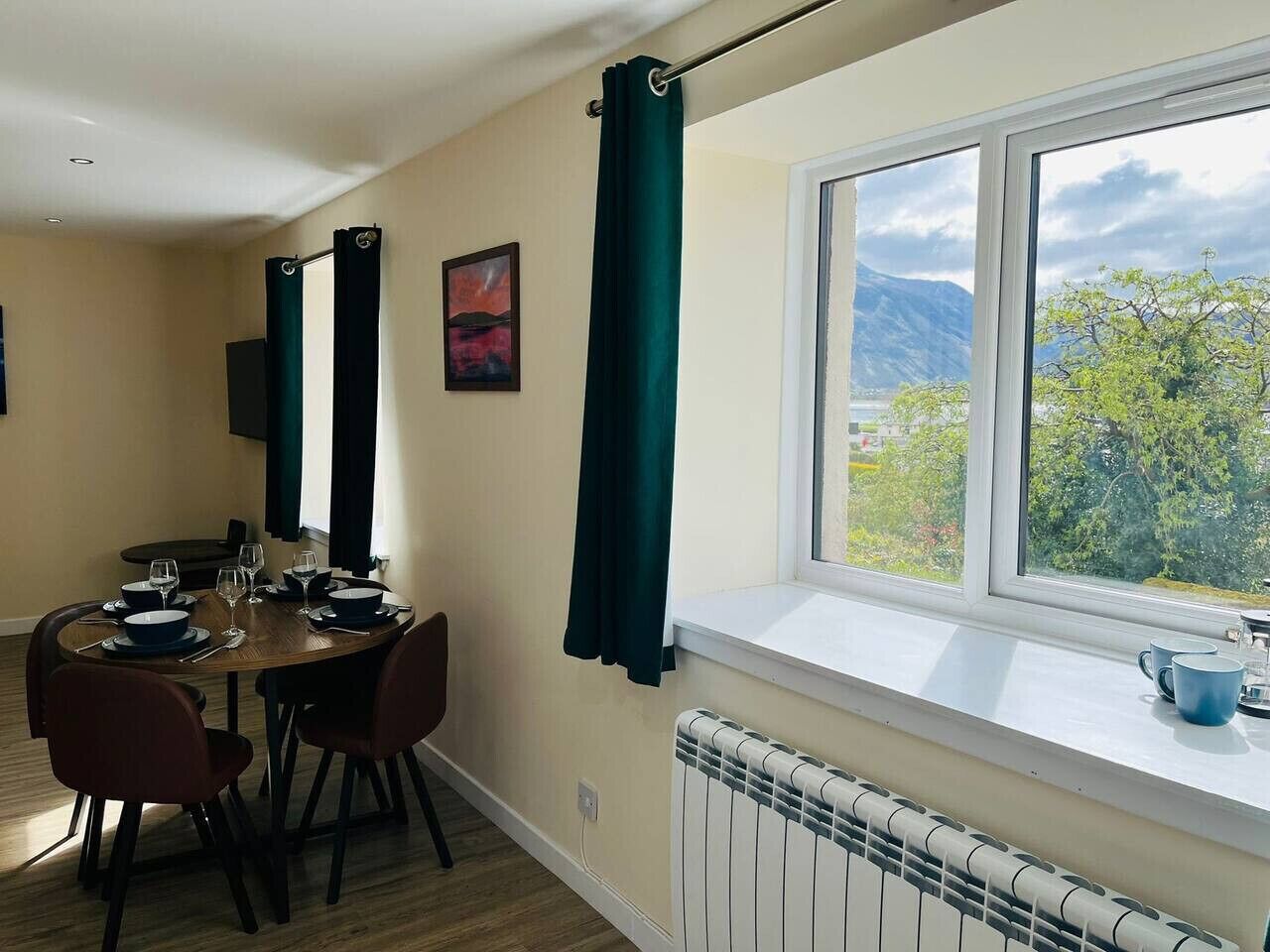 Un coin repas avec une table dressée pour quatre personnes, une fenêtre donnant sur un paysage verdoyant et des montagnes, et un radiateur placé sous la fenêtre.
