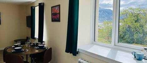 Un coin repas avec une table dressée pour quatre personnes, une fenêtre donnant sur un paysage verdoyant et des montagnes, et un radiateur placé sous la fenêtre.