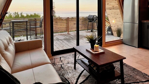 A modern interior with a large sliding glass door leading to an outdoor deck, a sofa, a coffee table, and a kitchen area. - Summit A-Frame (Two Harbors)