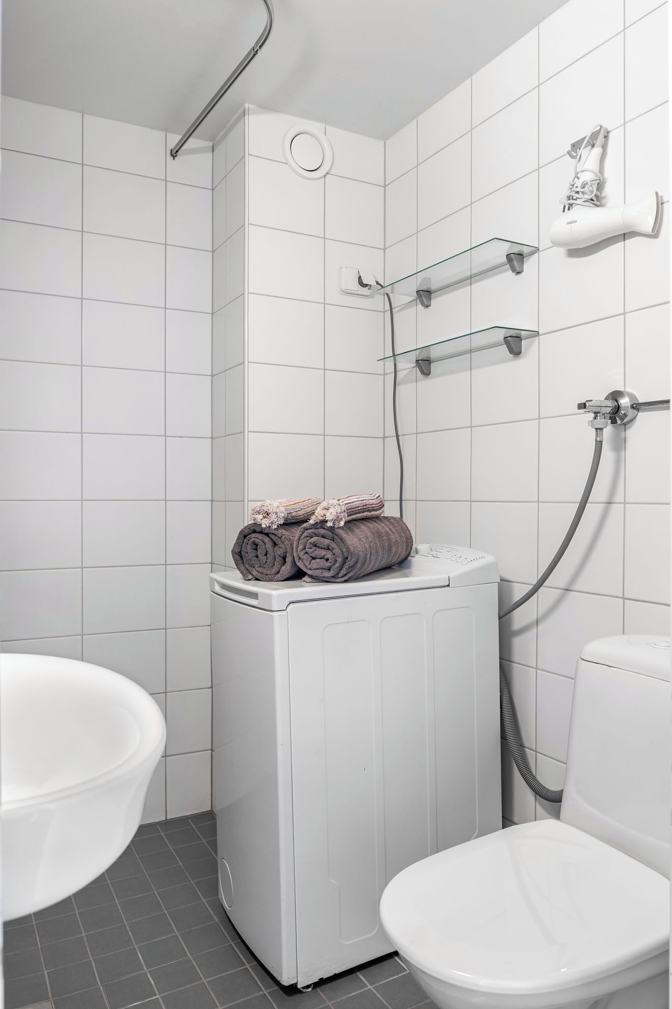 A bathroom with a white tiled wall, a white toilet, a white washing machine, and a shower with a glass shelf.