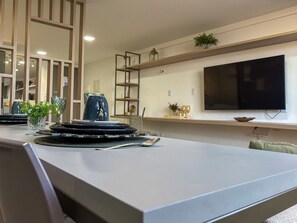 Un comedor moderno con una larga mesa dispuesta para una comida, un televisor montado en la pared y un estante decorativo con plantas y cristalería.