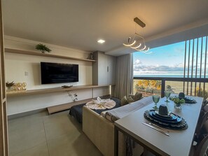 A modern living room with a flat-screen TV, a dining area with a table set for two, and a balcony with a view of the ocean. - FlatFM20 CABO BRANCO Oceanview by Almare (João Pessoa)
