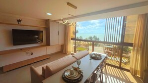 A modern living room with a flat-screen TV, a sofa, a dining table set with plates and a glass, and a balcony with a view of the ocean. - FlatFM20 CABO BRANCO Oceanview by Almare (João Pessoa)